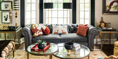 A gray tufted sofa with red pillows sits in a bright living room with glass tables, art, and decorative objects.
