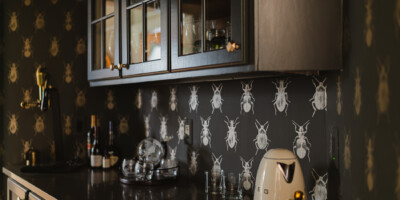 A kitchen counter with glassware, bottles, and appliances, set against wallpaper with a repeating beetle pattern.