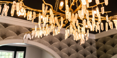 A gold chandelier with crystal accents hangs from the ceiling in a room with patterned gray wallpaper and arched doorways.