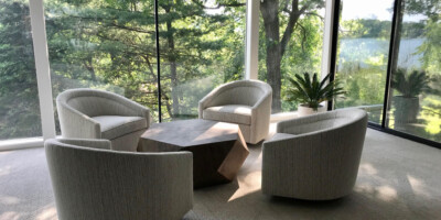 Four modern armchairs surround a geometric coffee table in a glass-walled room overlooking trees and greenery.